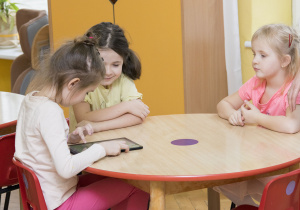 trzy dziewczynki siedzą przy stoliku i bawią się tabletem